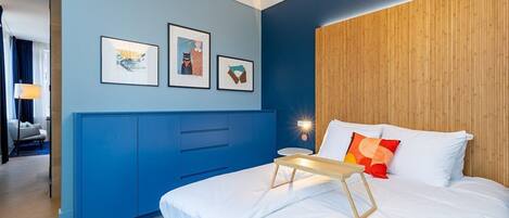 A modern hotel room with a blue accent wall, a wooden headboard, a bed with white bedding, and a small bedside table.