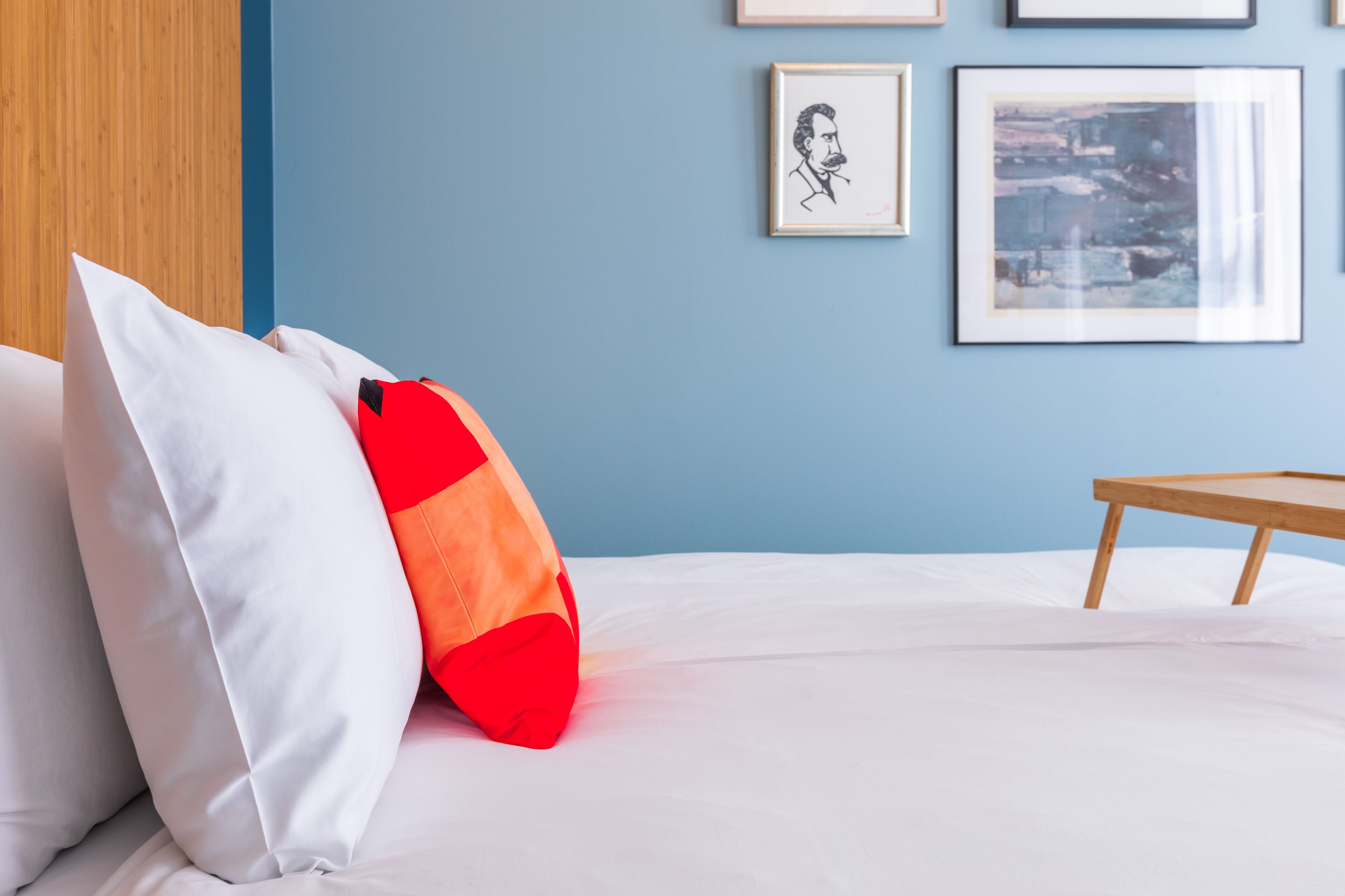 A neatly made bed with white and red pillows, a wooden bedside table, and framed artwork on the wall.