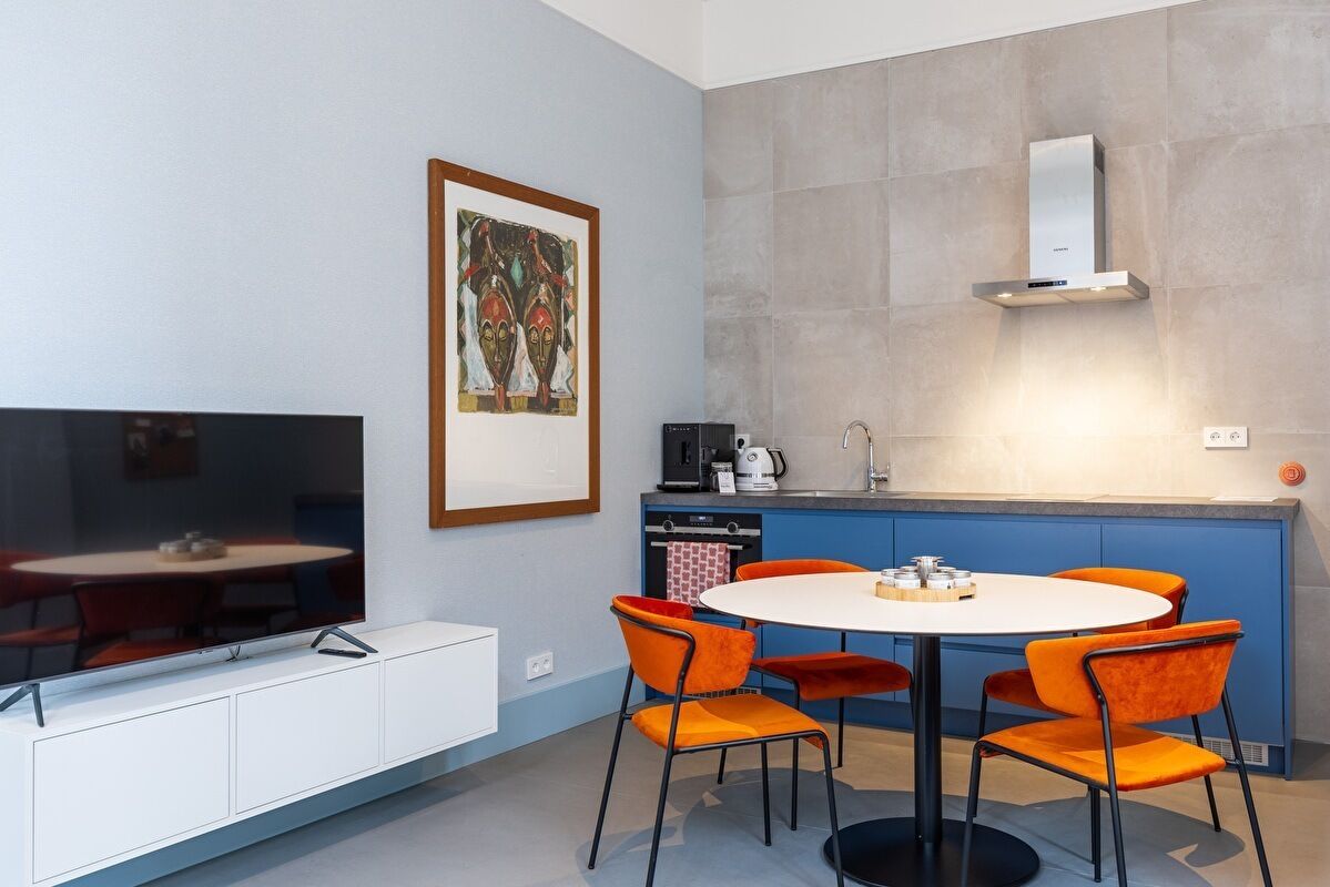 Una cocina moderna con horno empotrado, cafetera y microondas. Hay una mesa de comedor con sillas anaranjadas y mantel blanco. Una obra de arte enmarcada está en la pared sobre la mesa.