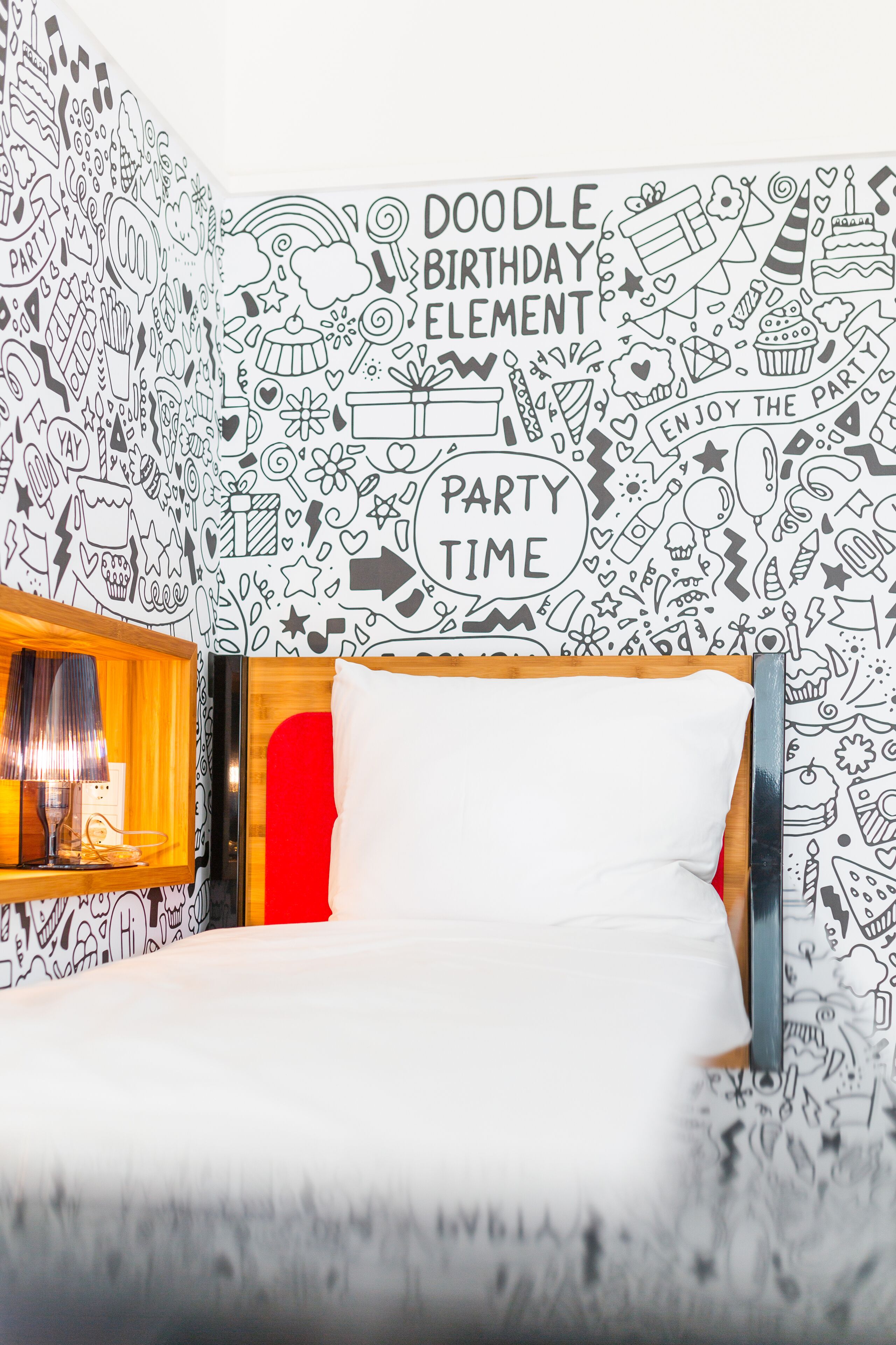A room with a bed, a nightstand, and a wall decorated with doodle birthday elements.