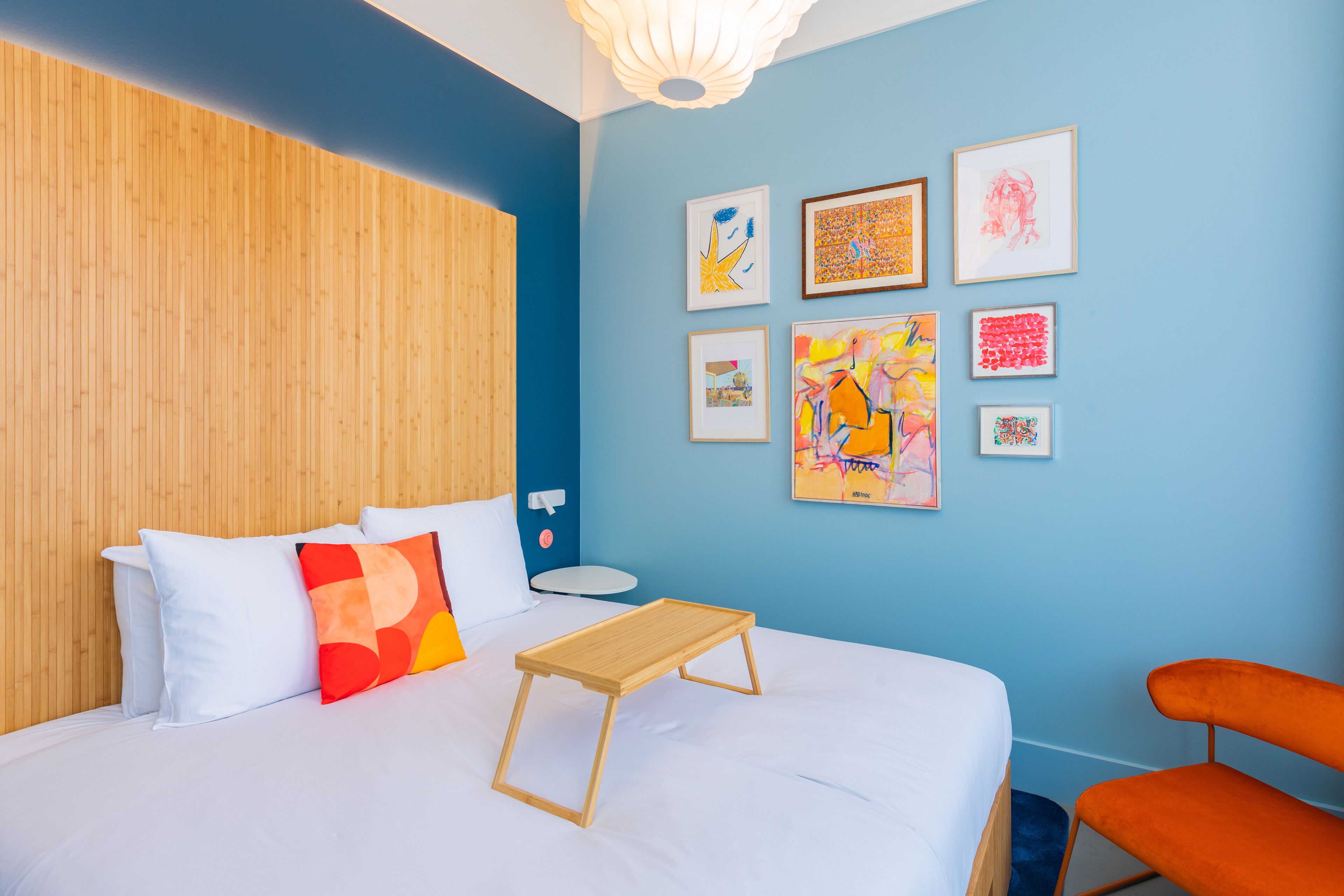 A room with a bed, a wooden headboard, a small table, an orange chair, and several framed artworks on the wall.