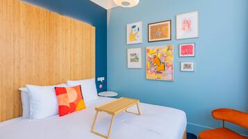 A room with a bed, a wooden headboard, a small table, an orange chair, and several framed artworks on the wall.