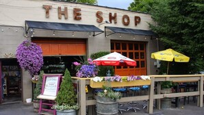 A shop with a sign reading "THE SHOP" above the entrance. There are outdoor tables with umbrellas, potted plants, and a signboard. - Quaint South Hill Home w/ Luxury King Bed & Wifi! (Spokane)
