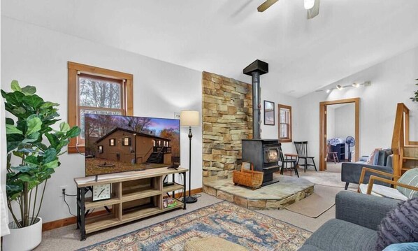 A cozy living room with a stone fireplace, a television, and a large plant. - River Access Cabin with Hot Tub, Fire Pit & WiFi! (Berkeley Springs)