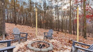 A forested area with Adirondack chairs arranged around a fire pit.