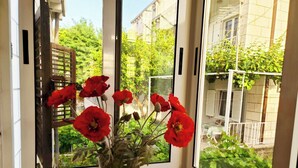 A window with a view of a garden and buildings, a vase with red flowers, and a wooden table.