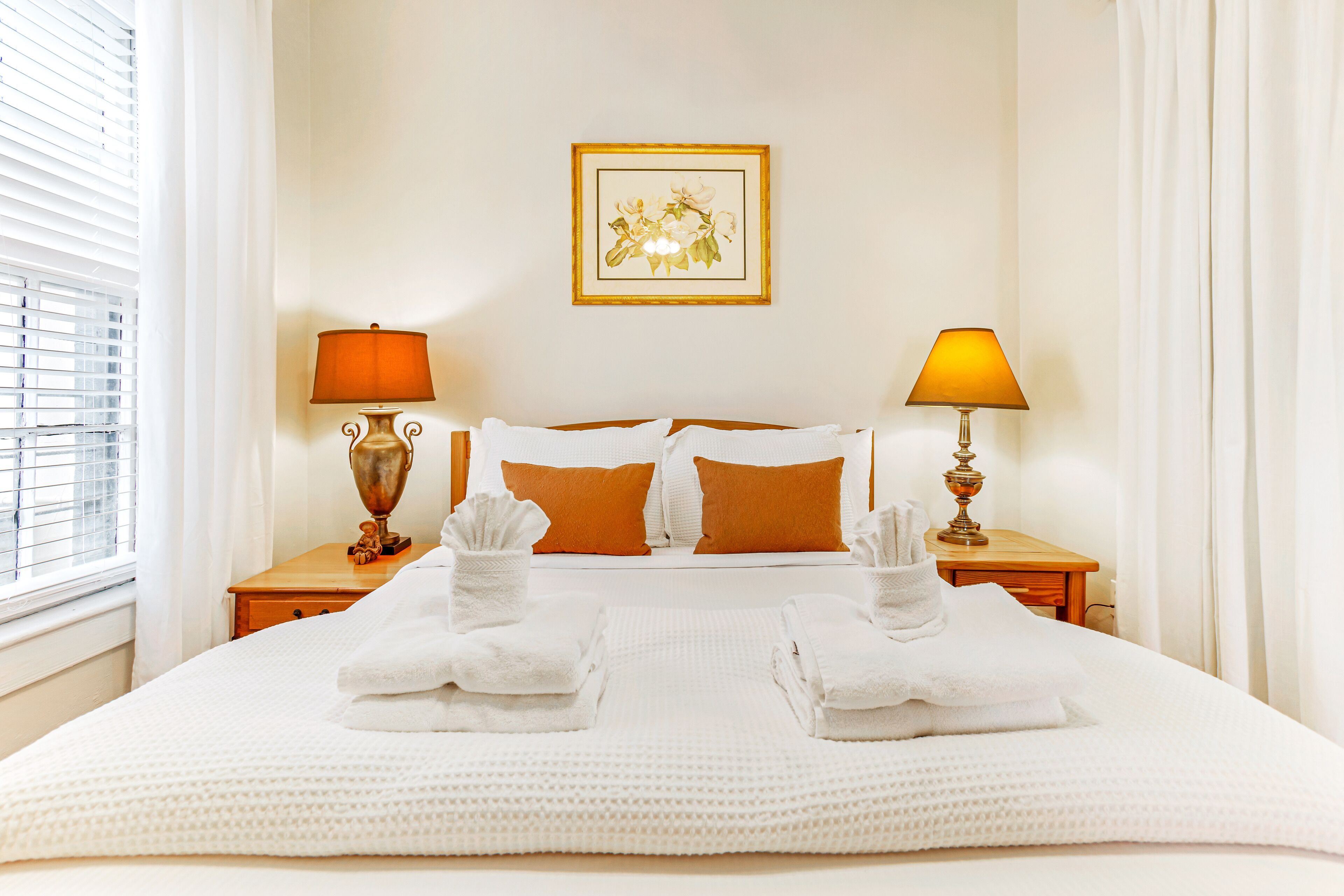 Cama bem arrumada com lençóis brancos, duas luminárias de cabeceira e uma obra de arte floral emoldurada na parede.