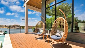 Una terraza de madera con dos sillas de mimbre y una mesa pequeña, con vistas a un cuerpo de agua y edificios.