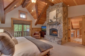 A spacious bedroom with a large bed, a stone fireplace, and a balcony with a wooden railing.
