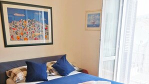 A neatly made bed with a blue bedspread and two towels, a framed abstract painting above the bed, and a window with a view of buildings. - Insideout Apartment (Dubrovnik)