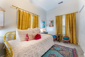 A bedroom with a yellow metal bed frame, a white bedspread, and yellow curtains. There is a small table with a lamp and a chair.