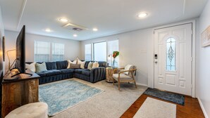 Un salón con un sofá modular, una mesa de centro y un televisor. Hay dos ventanas con persianas, una puerta con panel de vidrio y una alfombra en el suelo.
