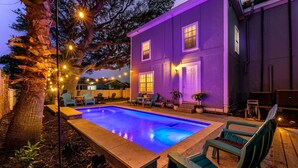 Una piscina con cubierta de madera, rodeada de mobiliario exterior y un edificio morado.
