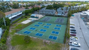 Vue aérienne d’un complexe de courts de tennis comprenant plusieurs terrains, entouré de bâtiments résidentiels et d’un parking.
