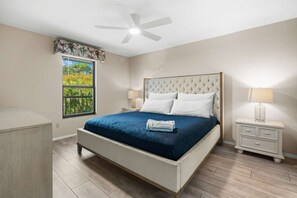Une chambre à coucher comprenant un lit, deux tables de chevet, un ventilateur de plafond, une fenêtre avec vue et une petite table de chevet avec une lampe.