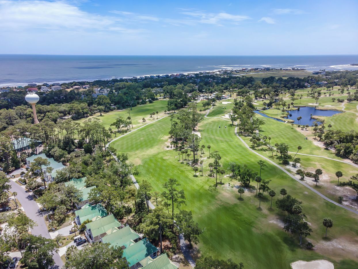 An aerial view of a golf course with a large body of water in the background, surrounded by residential areas and greenery.