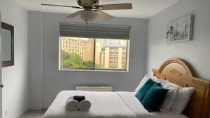 A hotel room with a bed, a ceiling fan, a window with a view of buildings, and a painting on the wall. - Heart of ATL Downtown  1B1B  City View  Cozy Condo (Atlanta)