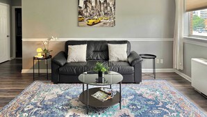 Un salon avec un canapé en cuir noir, une table basse en verre, un tapis à motifs, un tableau représentant un paysage urbain accroché au mur et une fenêtre avec des stores.
