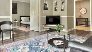 Un salon moderne avec un téléviseur à écran plat, une table basse avec des magazines, un tapis à motifs et un lit en arrière-plan.