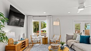 En moderne stue med et fladskærms-tv, to lænestole, en sofa, et sofabord og et stort vindue med gardiner.