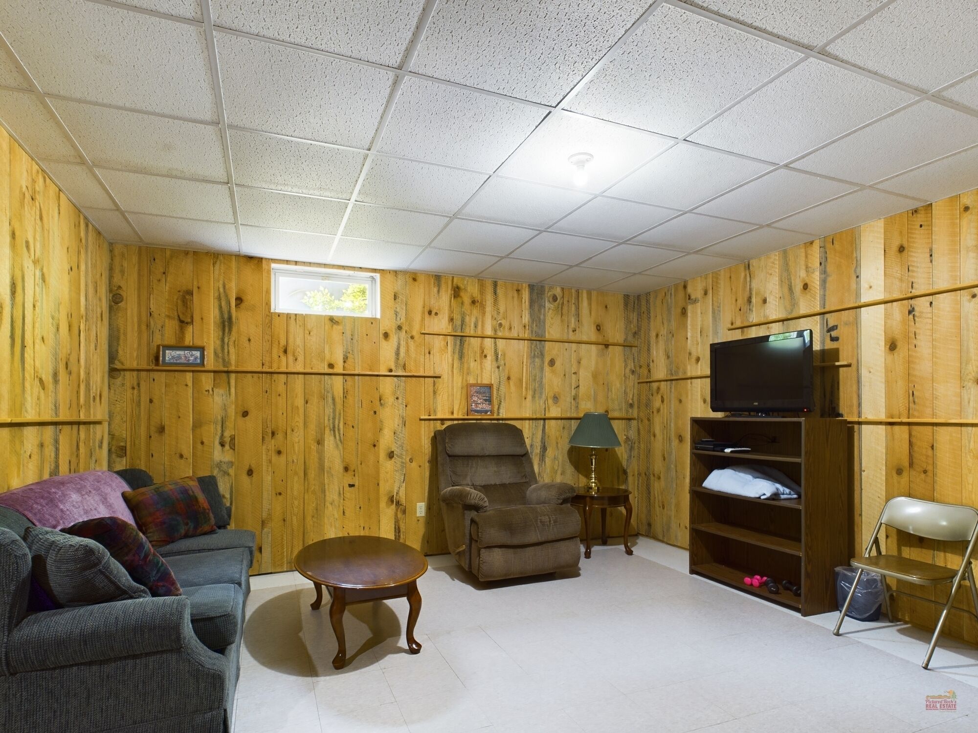 Un salón con paredes de madera, un televisor sobre un mueble bajo, un sofá, una silla, una mesita y una estantería.