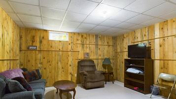 Un salón con paredes de madera, un televisor sobre un mueble bajo, un sofá, una silla, una mesita y una estantería.