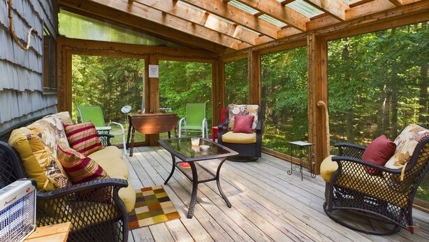 A covered outdoor seating area with wicker furniture, a glass table, and a view of the surrounding forest. - Spacious UP Retreat with Sauna & Direct Trail Access (Wetmore)