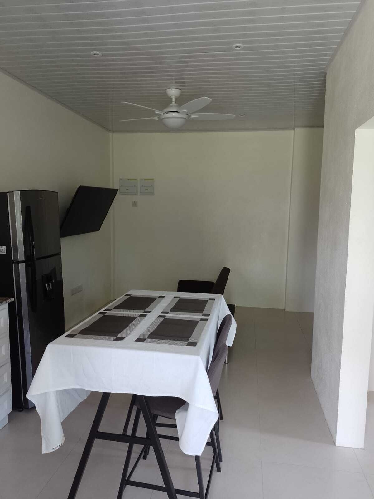 A compact room with a dining table, chairs, a ceiling fan, and a refrigerator.