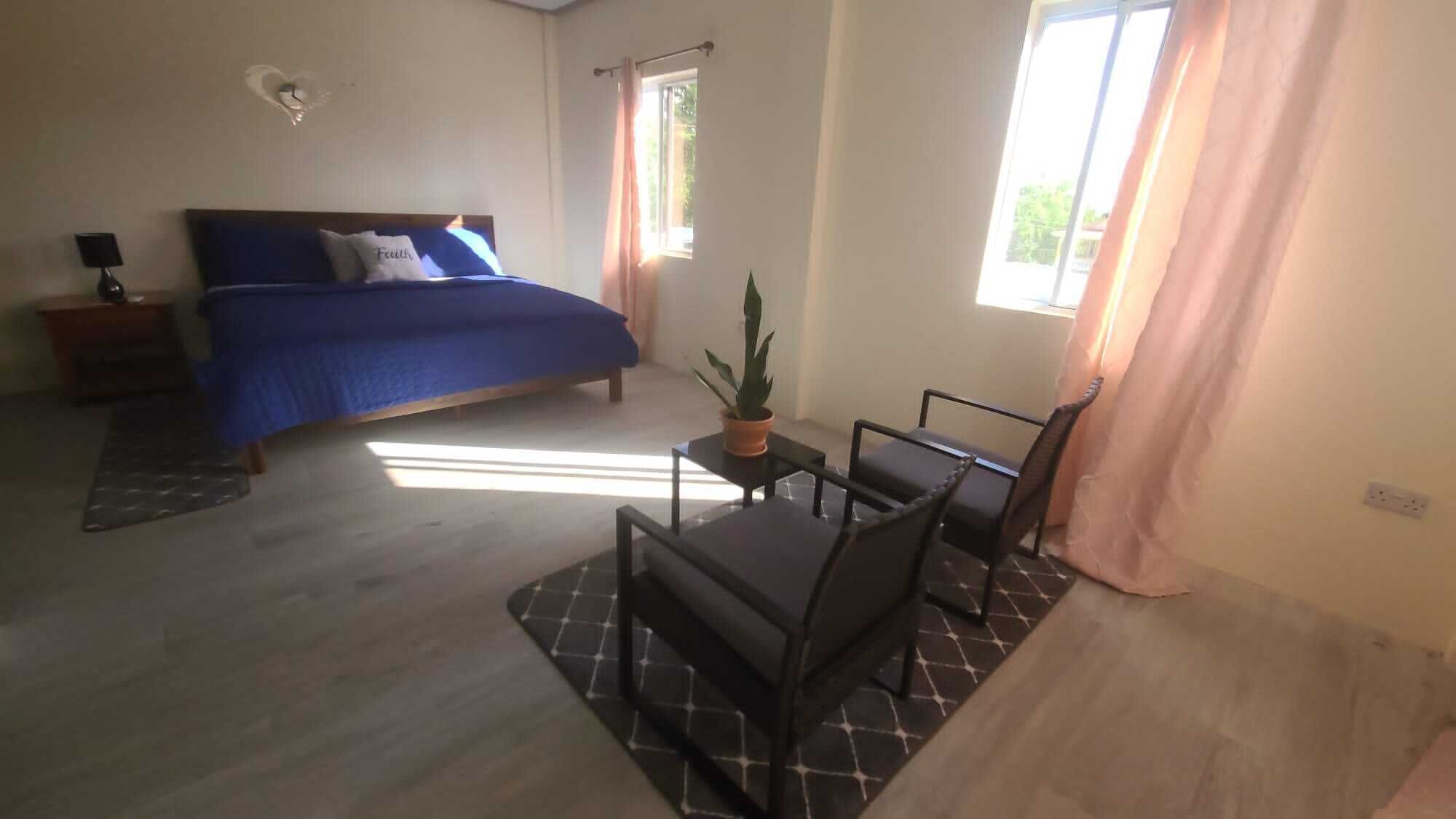 A bedroom with a bed, a small table with a potted plant, two chairs, a nightstand, and a window with curtains.