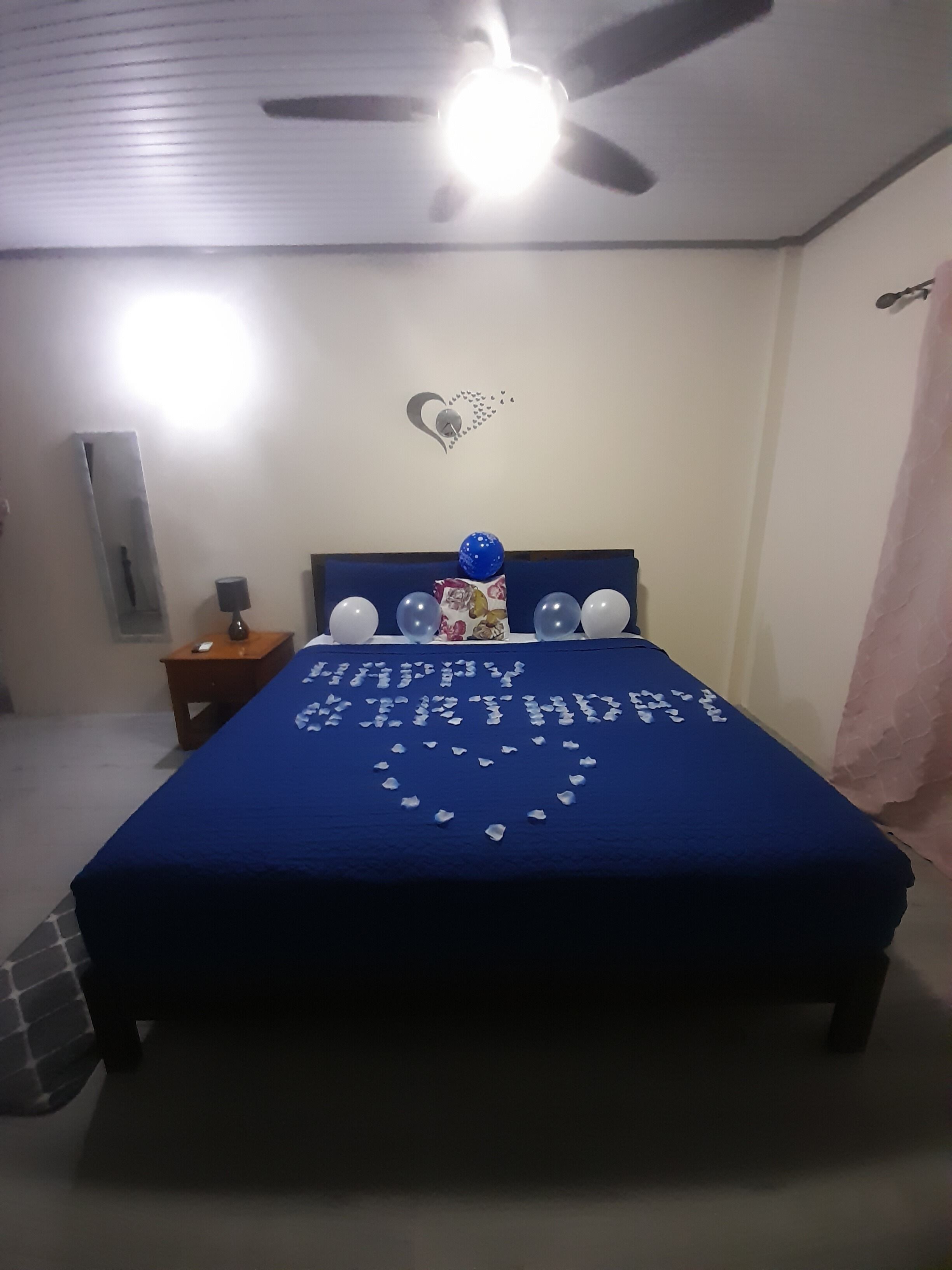 Un dormitorio con cama, ventilador de techo, decoración en la pared y mesita de noche con lámpara.