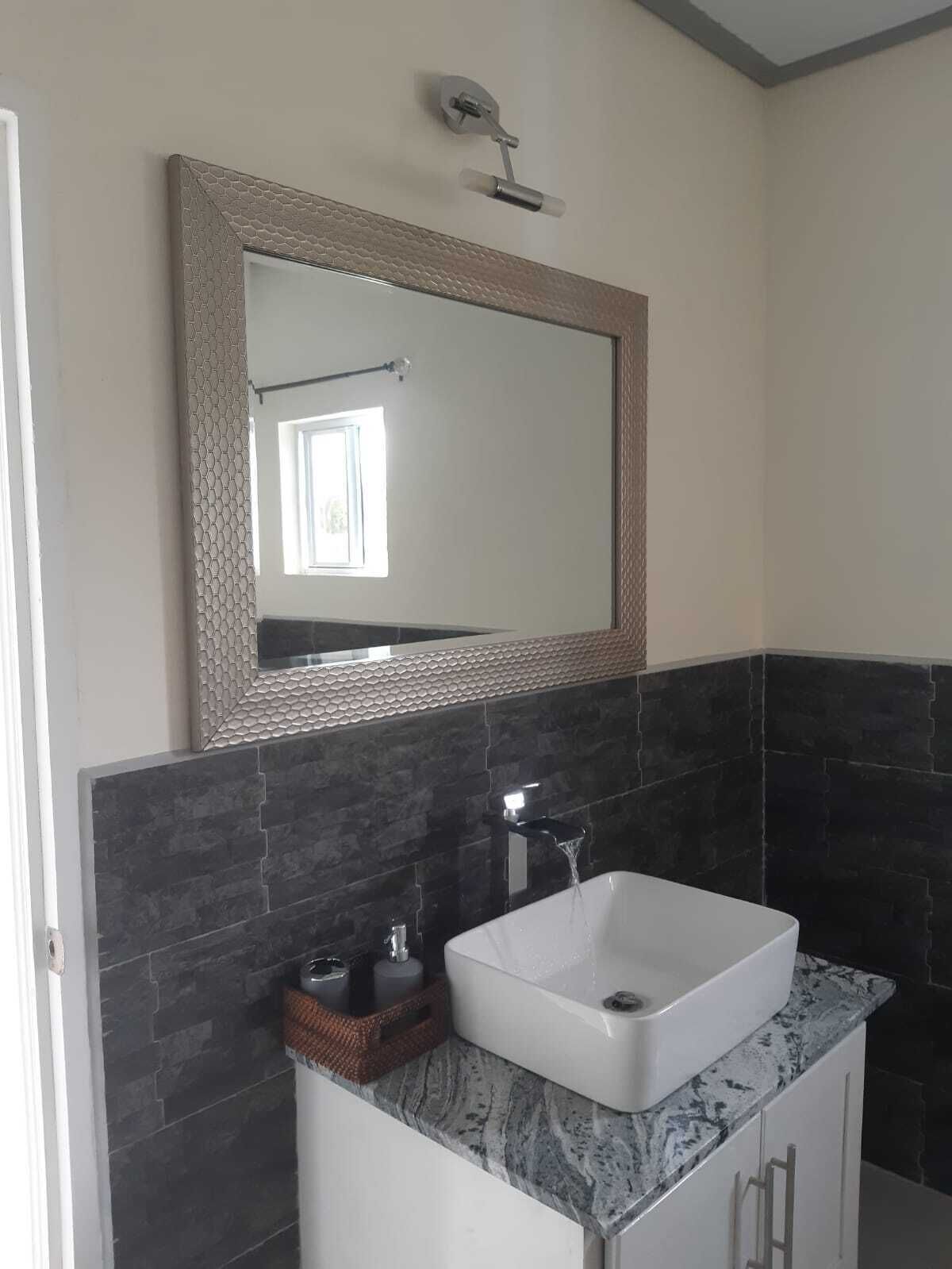 A bathroom with a white sink, dark tiled walls, a mirror with a textured frame, and a small window.