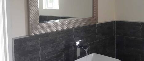 A bathroom with a white sink, dark tiled walls, a mirror with a textured frame, and a small window.