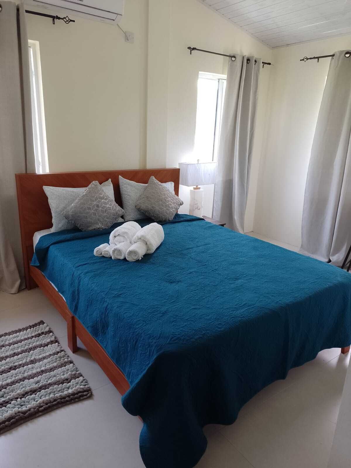 A neatly made bed with a blue bedspread and white pillows, a wooden headboard, and a white bedside table with a lamp.