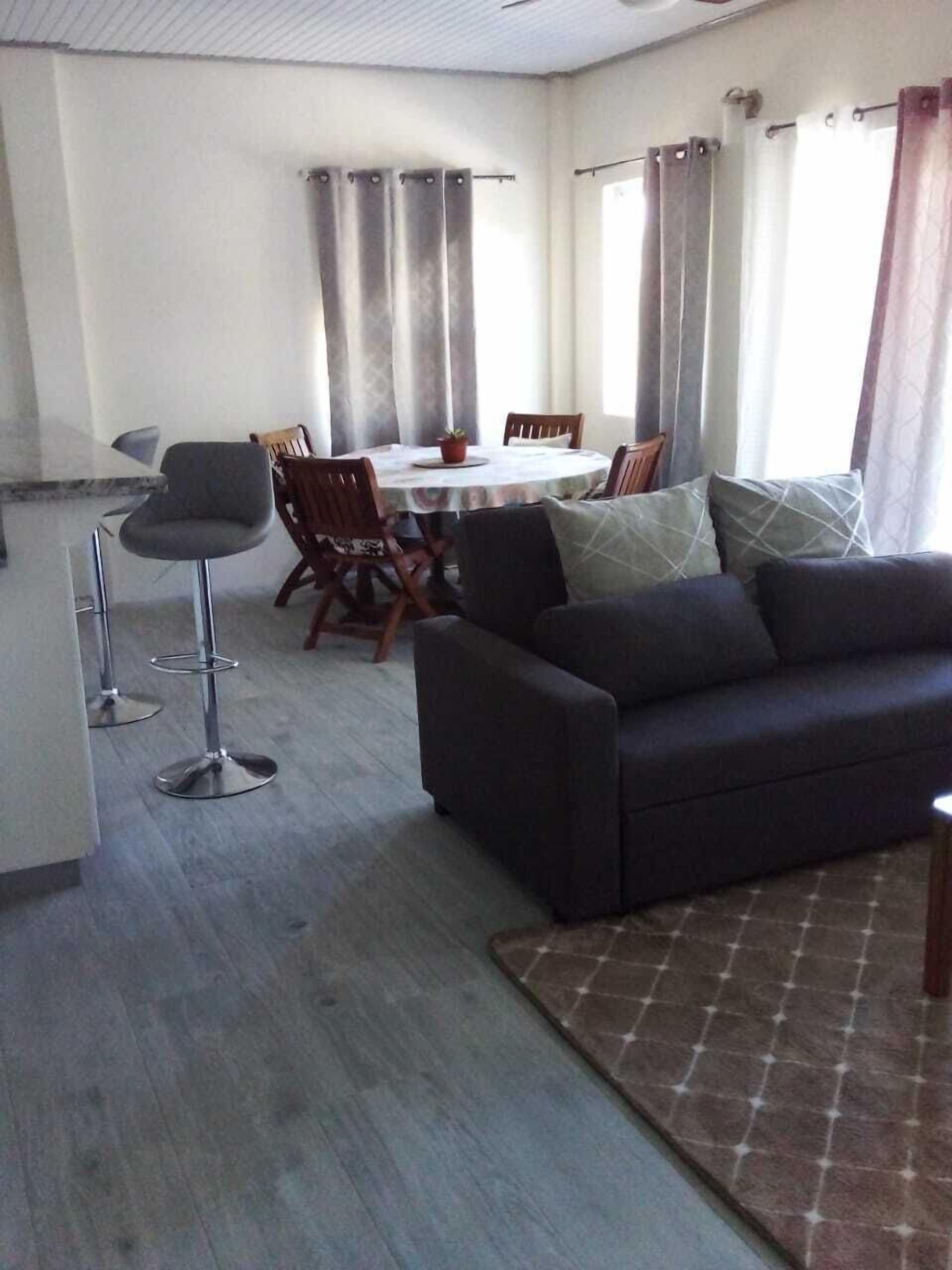 A living room with a dark sofa, a dining table with chairs, and a kitchen area with a white countertop and bar stools.