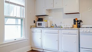 Une cuisine avec des meubles blancs, un four à micro-ondes, une cuisinière et une fenêtre avec des stores.
