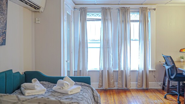 Une chambre d’hôtel avec un lit, un bureau et une chaise. Il y a une fenêtre avec des rideaux, un appareil de climatisation et un luminaire fixé au mur.