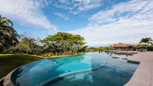 A clear swimming pool with a curved edge, surrounded by a well-maintained lawn and trees. - Hilltop condo with beautiful ocean view! (Tarcoles)