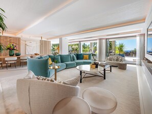 A spacious living area with a large sofa, a coffee table, and a TV. There is a balcony with a view of the ocean.
