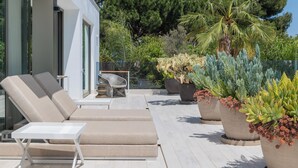 Un espace de détente extérieur moderne, avec des fauteuils beiges et une table blanche, entouré de grands pots de plantes et d’une balustrade en verre donnant sur un jardin luxuriant.