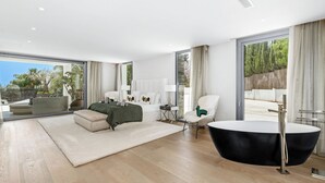 Un vaste espace de vie comprenant un grand canapé, une table basse ronde noire et un fauteuil blanc. Une grande fenêtre offre une vue sur la verdure environnante.