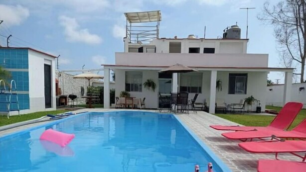 A swimming pool with lounge chairs, a covered patio, and a modern building with a flat roof.