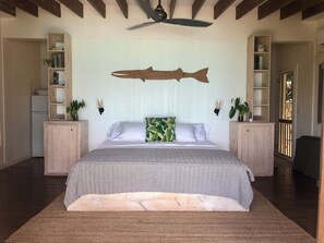 Ein Schlafzimmer mit einem großen Bett, einem Holz-Kopfteil, zwei Nachttischen mit Lampen, einem Deckenventilator und integrierten Regalen mit Büchern und Pflanzen.