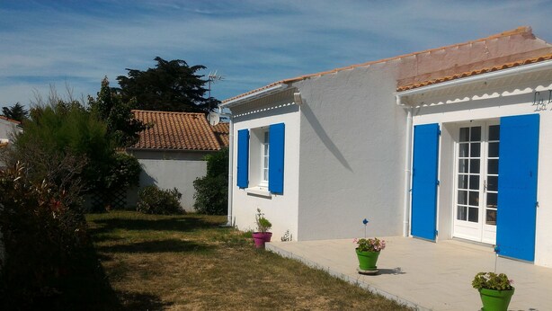 Un edificio de paredes blancas y contraventanas azules, con techo de tejas y un pequeño jardín con plantas en macetas.