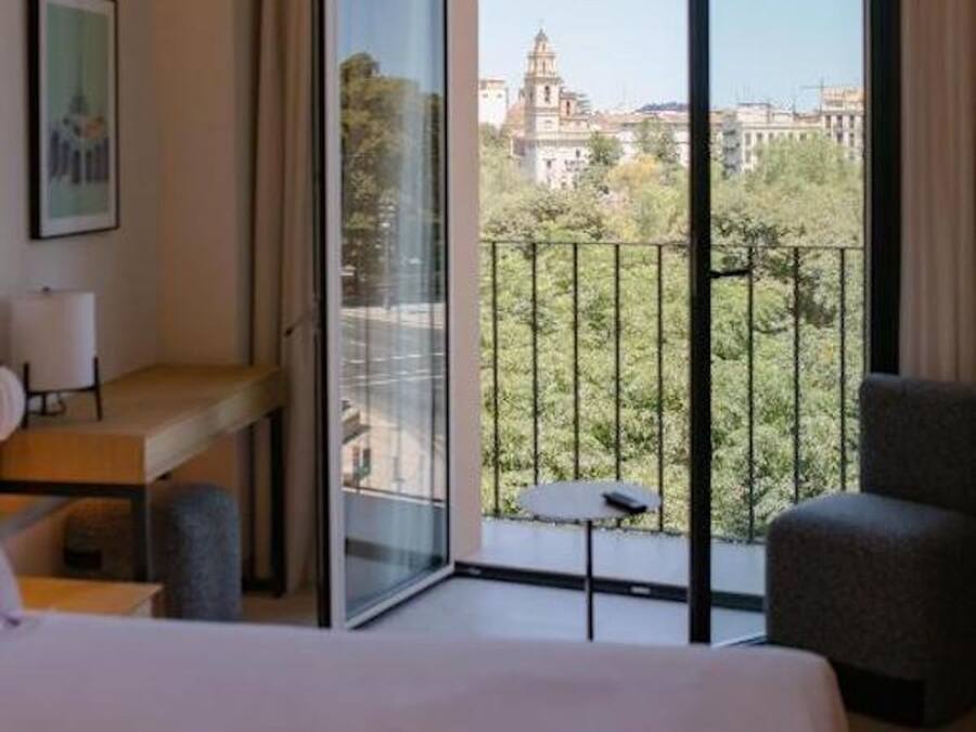 A hotel room with a bed, a desk, a chair, and a balcony with a view of a cityscape.