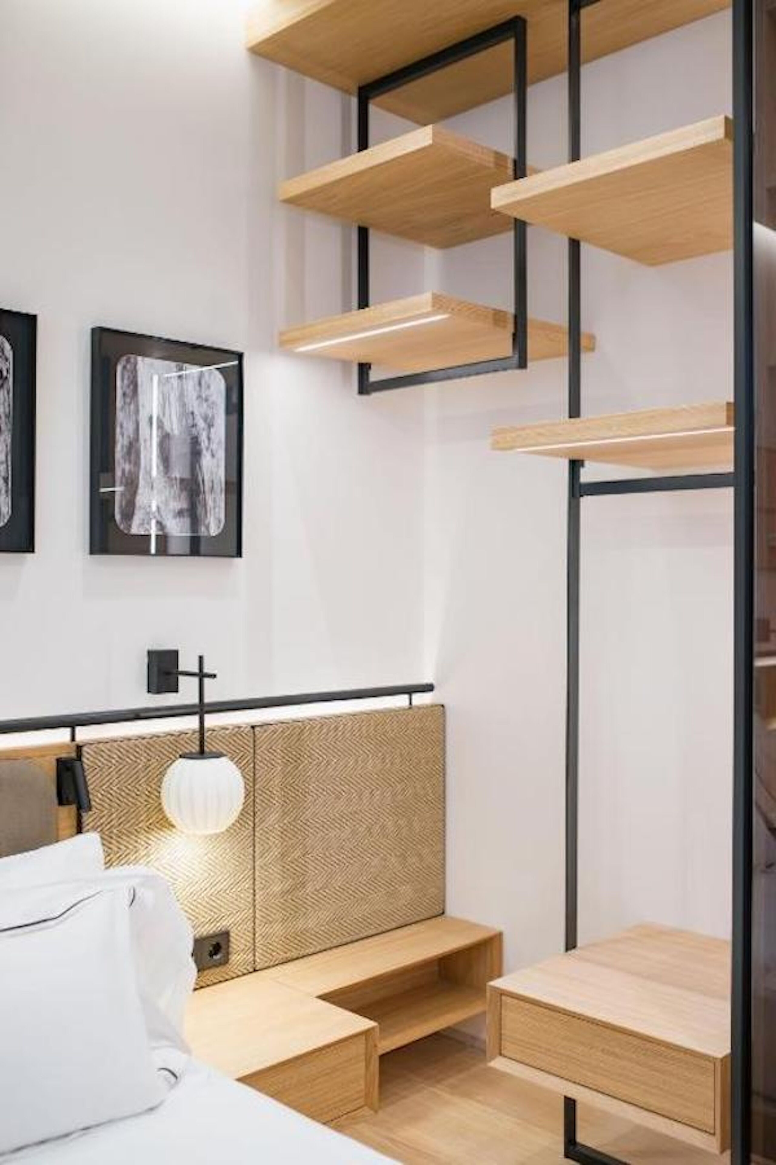 Una habitación de hotel moderna con una cama, un banco de madera, un estante en la pared y una lámpara de pared.