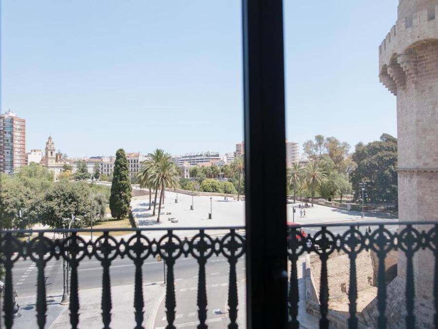 Vista desde un balcón con barandilla de hierro forjado, sobre un paisaje urbano con edificios modernos, palmeras y una torre de piedra histórica.