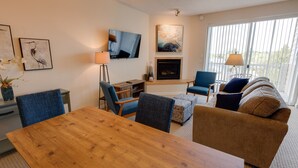 A living room with a wooden dining table, blue and brown upholstered chairs, a blue armchair, a sofa, a fireplace, and a TV mounted on the wall. - Calliope's Seaside Sanctuary Condominium - Oyhut Bay Seaside Village (Ocean Shores)