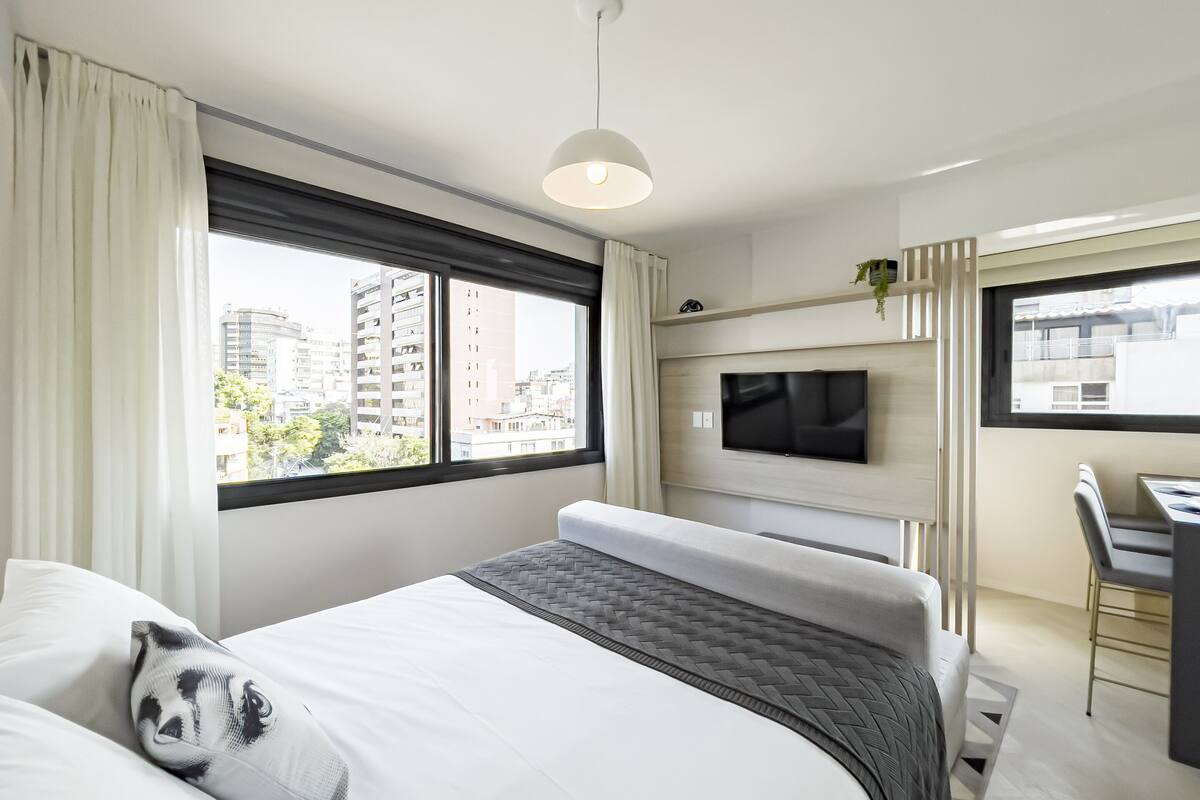 A modern hotel room with a large bed, a TV mounted on the wall, a desk with a chair, and a view of the city through the window.
