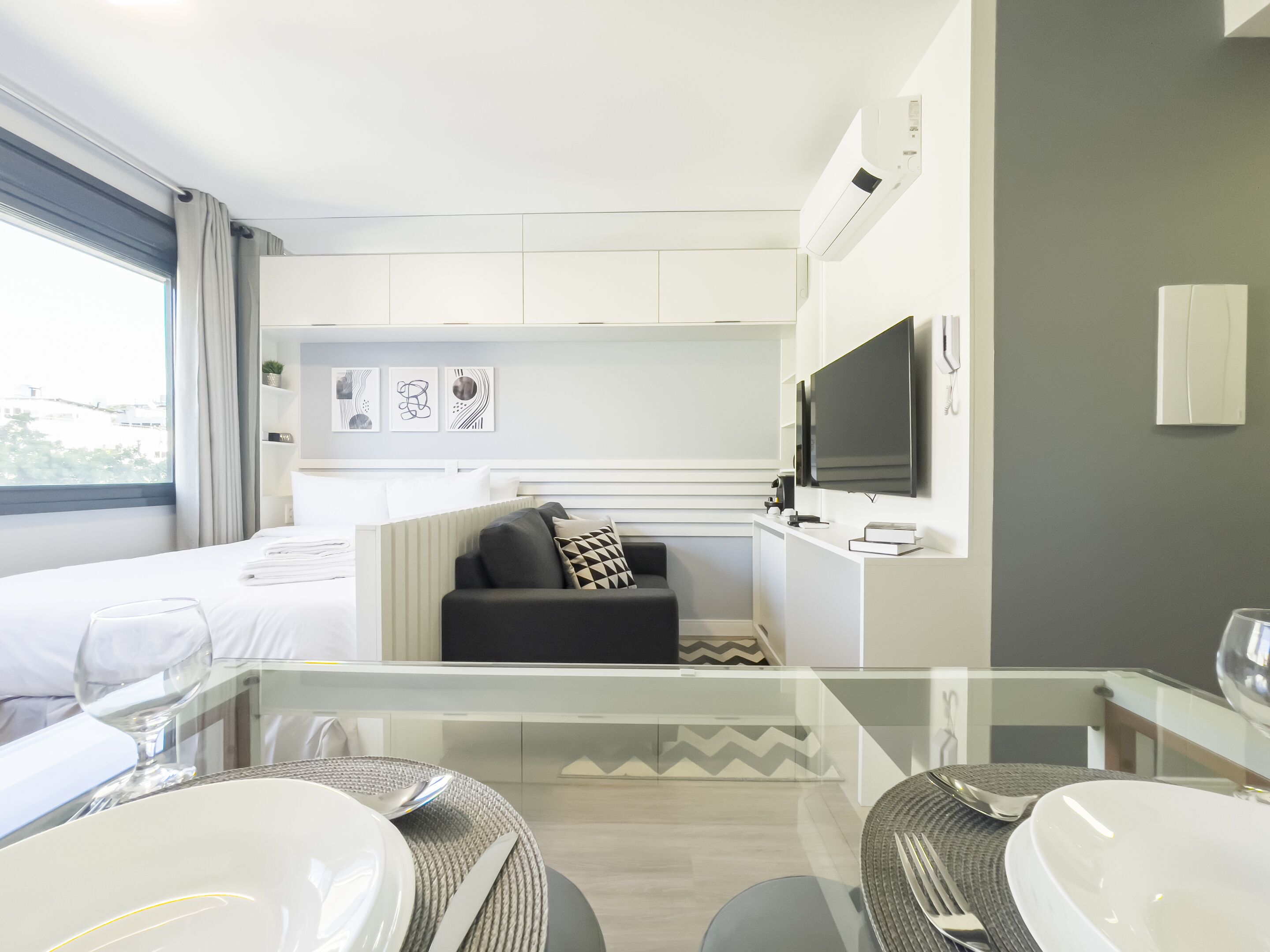 A modern hotel room with a glass dining table, a sofa, a flat-screen TV, and artwork on the wall.