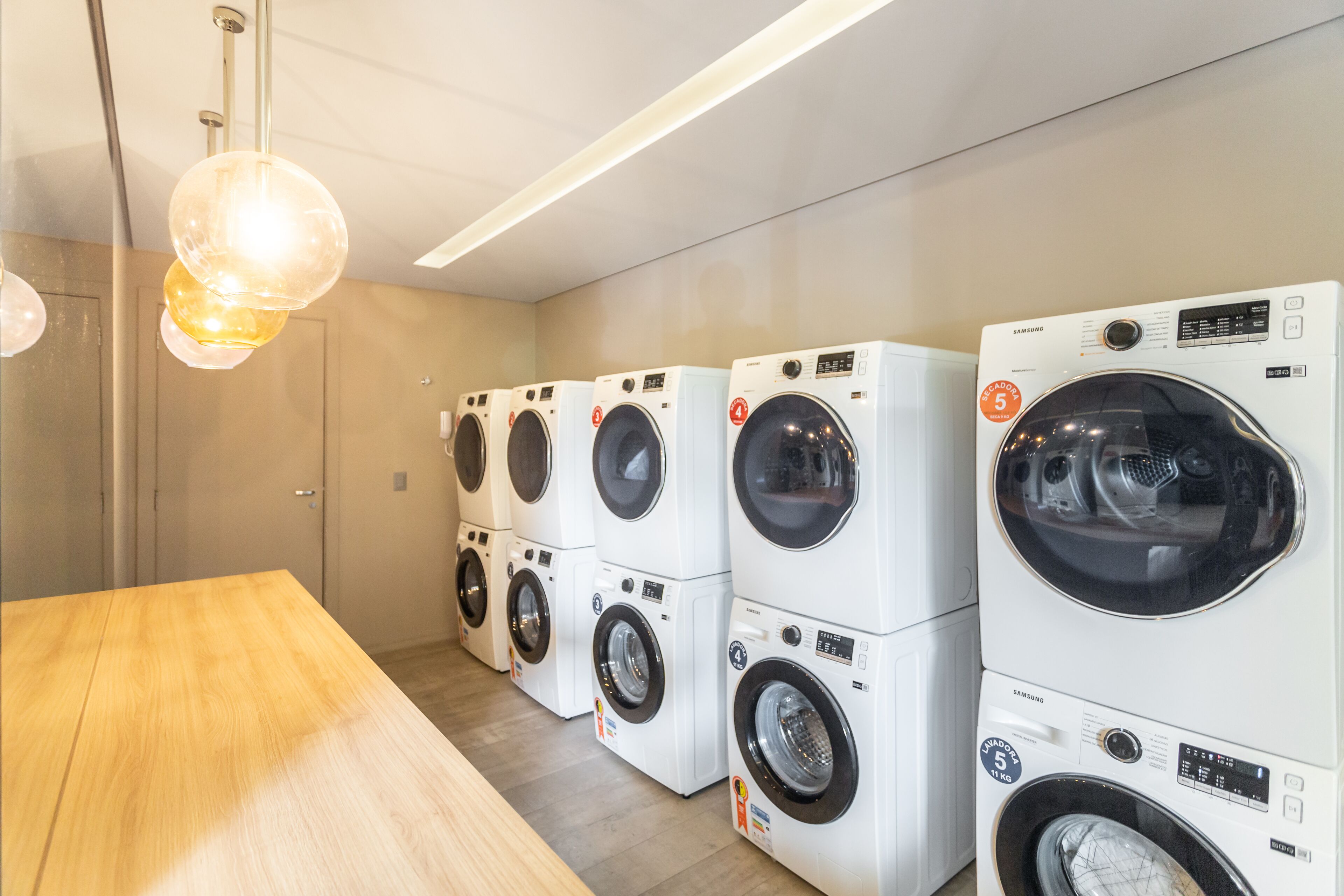 A row of Samsung washing machines in a laundry room with a wooden table and a hanging light fixture.